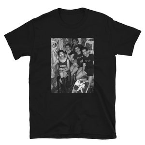 May include: Black and white photograph of five people standing in a room. The people are wearing t-shirts with the words "DEVO" and "REVERSE EVOLUTION" printed on them.