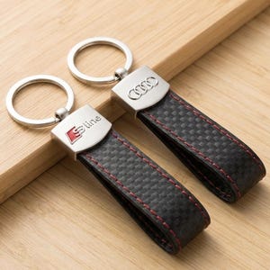 May include: Two keyrings with silver rings and black carbon fibre straps. One keyring has the Audi logo, the other has the S line logo. Both have red stitching and are on a wooden surface.