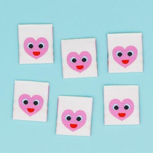 May include: Six white fabric patches with a pink heart-shaped face design, featuring black eyes and a red smiling mouth. The patches are arranged on a light blue background.