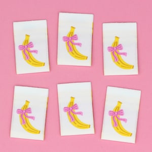 Puede incluir: Seis etiquetas de tela blanca con un diseño de plátano amarillo y un lazo rosa. Los plátanos tienen contornos marrones y están sobre un fondo rosa. Las etiquetas son rectangulares y tienen un borde cosido.