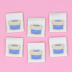 May include: Six square white fabric labels, each featuring an embroidered illustration of a beige tub with a blue label and the text HUMMUS in blue letters. The tub lid is light blue with three small pink hearts centred on the top surface.