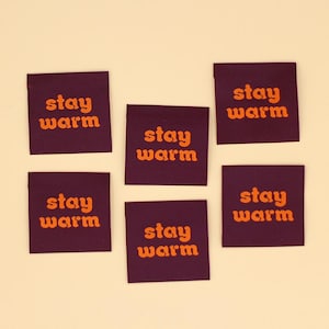 May include: Six square, dark purple fabric labels with the words "stay warm" in orange lettering. The labels are arranged on a light beige background. The text is in a retro font.