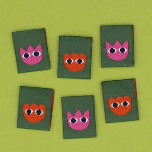 Puede incluir: Seis parches de tela verde con un diseño de flor rosa o naranja y ojos blancos. Los parches tienen un borde rojo y están dispuestos sobre un fondo verde claro.
