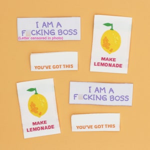 May include: White fabric labels with text and illustrations on an orange background. The labels read "I AM A F***ING BOSS", "YOU'VE GOT THIS", and "MAKE LEMONADE" with a yellow lemon illustration.