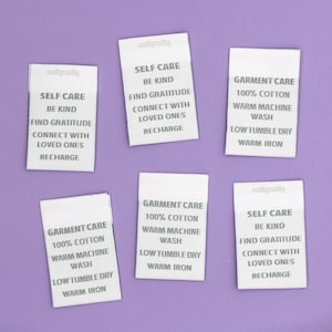 May include: Six white fabric labels on a purple backdrop. The labels display text including "Self Care, Be Kind, Find Gratitude" and "Garment Care, 100% Cotton, Warm Machine Wash". The brand name "mathymatly" is also visible.
