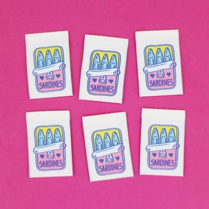 Op de afbeelding: Zes witte stoffen labels met een cartoon sardineblikje ontwerp. Elk label heeft een geel en blauw sardineblikje illustratie met het woord "SARDINES" en een hart symbool. De achtergrond is felroze.