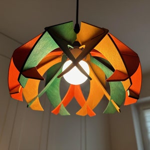 Op de afbeelding: Een kleurrijke hanglamp met een geometrisch ontwerp. De houten kap heeft oranje, groene en gele panelen. Een heldere gloeilamp is in het midden zichtbaar en verlicht de kamer. De armatuur hangt aan het plafond.