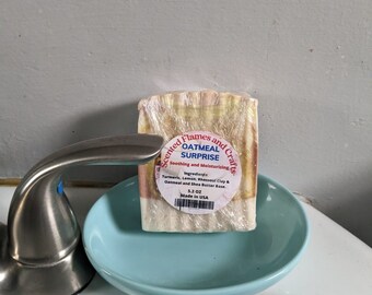 Puede incluir: Una barra de jabón de avena envuelta en plástico y colocada en un plato azul. El jabón está etiquetado como "Scented Flames and Crafts Oatmeal Surprise" y enumera los ingredientes como "Cúrcuma, Limón, Arcilla Rhassoul & Avena y Base de Manteca de Karité." El jabón pesa 5,2 oz y está hecho en los EE. UU.