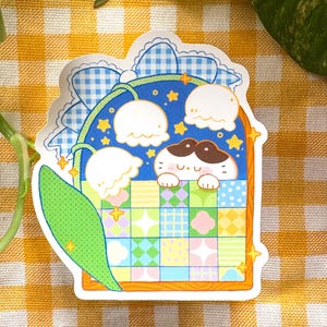 May include: A sticker featuring a cartoon cat peeking over a colorful patchwork quilt. The cat has a brown and white face and is surrounded by white flowers and stars. The sticker has a blue and white gingham border.