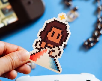 May include: A pixelated sticker of a person with brown hair and a halo, wearing a white robe with a red and blue pattern. The sticker is held in a hand over a white video game controller.