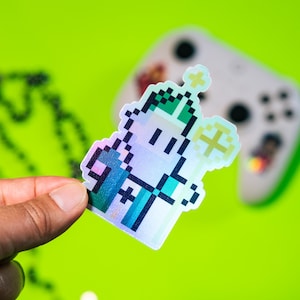 May include: A holographic sticker featuring a pixelated character with a green hat and a white body. The character is holding a golden staff with a star on top. The sticker is on a green background.