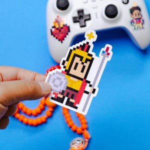 Könnte beinhalten: Ein pixeliger Aufkleber eines Cartoon-Ritters in gelber und roter Rüstung, der einen Schild und einen Stab hält. Der Aufkleber wird vor einem weißen Gamecontroller mit anderen Aufklebern und einer orangefarbenen Perlenkette gehalten. Der Hintergrund ist einfarbig blau.