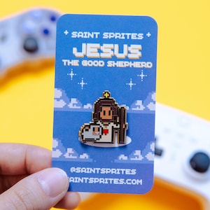 May include: A pixelated enamel pin featuring Jesus as the Good Shepherd, with a lamb, on a blue card. The card reads "SAINT SPRITES" and "JESUS THE GOOD SHEPHERD". The background is yellow.