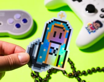 May include: A holographic sticker of a pixelated figure wearing a blue and white robe with a halo. The figure has brown skin and black eyes. The sticker is on a green surface with a video game controller and a beaded necklace.