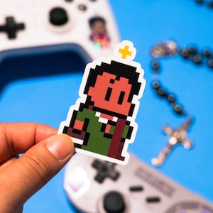 May include: A pixel art sticker of a person wearing a green robe and a red sash. The person has a halo above their head and is holding a book in their left hand. The sticker is on a blue background.