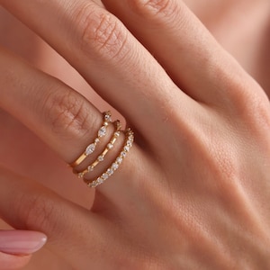 May include: A close-up of a hand wearing three gold rings. The rings are adorned with small, sparkling diamonds. The rings are stacked together on the finger. The background is blurred.