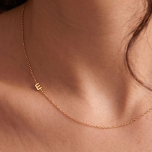 May include: A delicate gold-toned chain necklace featuring a single, small, uppercase letter E charm positioned off-center. The necklace has a fine link construction and rests against the collarbone, showcasing a simple and refined design.