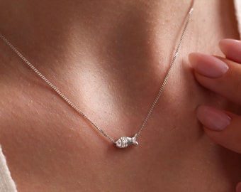 May include: A delicate silver necklace featuring a small fish pendant. The pendant is detailed with etched lines, and the chain is a fine, box-style link. The necklace is worn against a white top, creating a simple, elegant look.