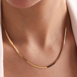 May include: A gold-colored, flat herringbone chain necklace. The necklace is worn around the neck, and the chain is visible against the skin and a white blazer.