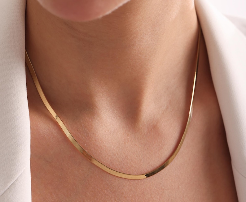 May include: A gold-colored, flat herringbone chain necklace. The necklace is worn around the neck, and the chain is visible against the skin and a white blazer.