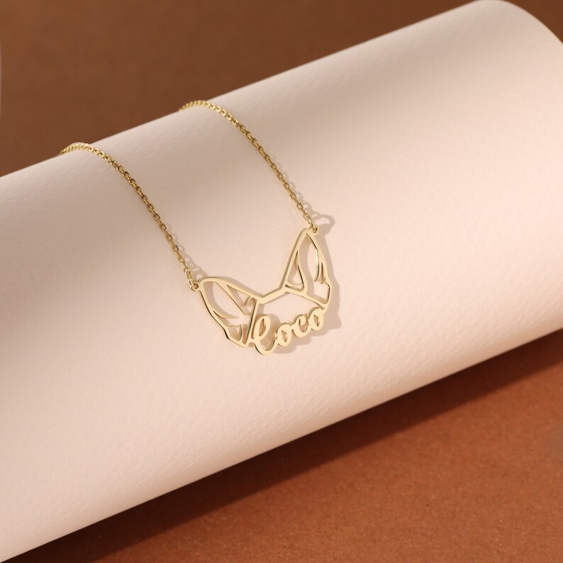 Puede incluir: Un collar de oro con una cadena delicada y un colgante con forma de cabeza de perro con el nombre "Coco" recortado.