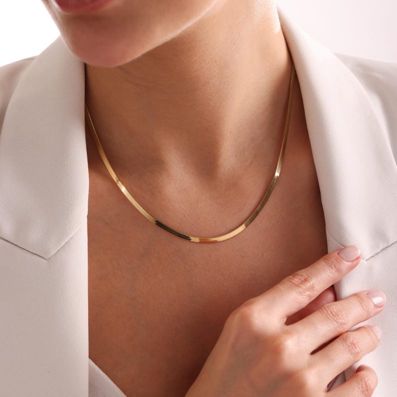 May include: A gold-colored herringbone chain necklace. The necklace is flat and lies smoothly against the skin. The image shows the necklace on a person wearing a white blazer.
