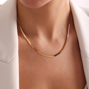May include: A gold-colored herringbone chain necklace. The necklace is flat and lies smoothly against the skin. The necklace is worn over a white blazer.