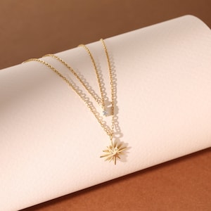 May include: A gold layered necklace with a starburst pendant and a small diamond accent. The necklace is on a white surface.