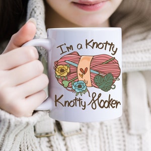 May include: White ceramic mug with a pink and green yarn ball, knitting needles, and the text "I'm a Knotty Knotty Hooker".