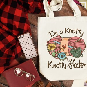 May include: Cream-colored tote bag with the text "I'm a Knotty Knotty Hooker" and a graphic of yarn, knitting needles, and flowers. The bag has a simple design with two handles. A red and black plaid shirt, a phone case, and a book are also in the image.