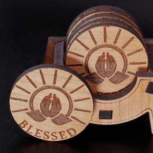 May include: Wooden coaster set with a holder. Each coaster features a sunburst design with praying hands and the word "BLESSED". The set is made of light brown wood with dark brown accents.