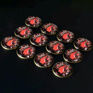 May include: A collection of twelve circular cabochon buttons featuring a graphic design of a red blood drop against a dark, splattered background. Each button has a smooth, domed glass surface set within a metallic gold-toned circular frame.