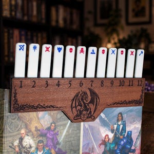 Puede incluir: Un accesorio de juego de mesa de madera oscura con un dragón grabado a láser y los números del 1 al 12. Doce marcadores rectangulares blancos con iconos azules y rojos, incluyendo calaveras, armas y símbolos, insertados en las ranuras.
