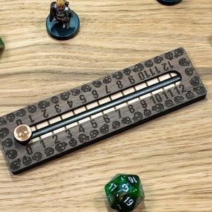 May include: A wooden score tracker for tabletop games, featuring a sliding marker, dice, and miniature figures. The tracker has engraved numbers and skull motifs. The dice are in various colours, including green and blue.