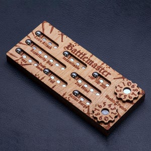 May include: A rectangular wooden game tracker labeled Battlemaster with engraved text and slots containing small metal ball bearings. It features multiple tracks for tracking game stats, including Extra Attack, Superiority Dice, and Health Points with two rotating gear dials.
