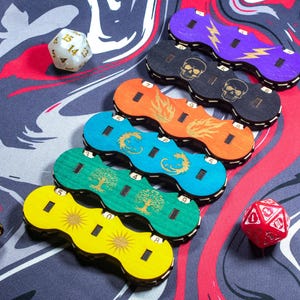 Puede incluir: Seis bandejas de dados de madera de colores con diferentes símbolos y números. Las bandejas están dispuestas en una fila de arriba a abajo, con la bandeja morada en la parte superior y la bandeja amarilla en la parte inferior. Cada bandeja tiene un símbolo diferente, incluyendo un rayo, una calavera, una llama, un dragón, un árbol y un sol.