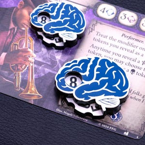 Op de afbeelding: Twee blauwe hersenvormige tokens met het witte nummer 8, op een spelkaart. De kaart heeft tekst en een afbeelding van een persoon die trompet speelt. Deze tokens zijn waarschijnlijk voor een bordspel.