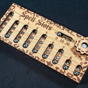 Peut inclure: Un plateau de suivi rectangulaire en bois clair avec le texte gravé Spell Slots et Sorcery Pts. Il comporte des fentes verticales avec de petites sphères métalliques sombres pour suivre les niveaux de sorts de un à huit, et un cadran en forme d'engrenage.
