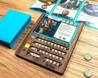 May include: A wooden card game organizer with slots for cards and tokens. A stack of blue cards sits to the left. Game cards and tokens are placed on the organizer. The organizer has the word "Actions" on it.