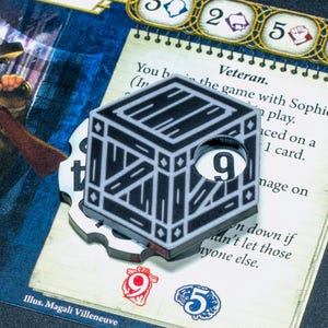 May include: A black and white square game token featuring a crate illustration with a circular cutout revealing the number 9. The token is layered over a game card with text including the word Veteran and various numerical icons.