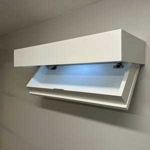 May include: A white, wall-mounted cabinet with a hinged, open door. The interior is illuminated with a soft, blue light. The cabinet is rectangular and appears to be made of wood or a similar material.