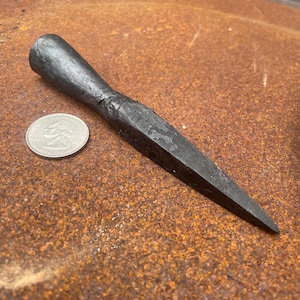 May include: A hand-forged dark gray metal tool with a tapered, pointed tip and a rounded, thicker handle end. A silver-colored United States quarter coin is placed next to the tool to provide a clear sense of scale for the object's size.