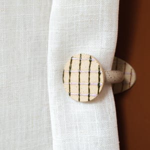 May include: A ceramic curtain tieback with a beige speckled finish. The circular tieback features a grid pattern of dark green and lavender lines. The tieback is holding a white linen curtain.