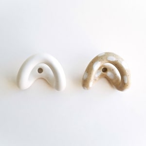 Pode incluir: Dois ganchos de parede de cerâmica em forma de arco. Um gancho é de cor branca mate sólida, enquanto o outro é de cor bege salpicada com manchas brancas pintadas. Cada peça apresenta um orifício central circular para a sua instalação.