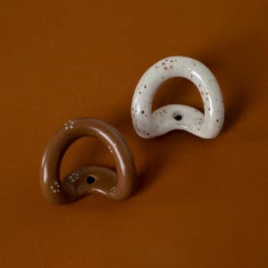 May include: Two D-shaped ceramic cabinet pulls, one brown and one white, with speckled patterns. Each pull has a central mounting hole. The pulls are set against a brown background.