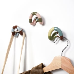 May include: Three ceramic wall hooks with a curved, looped design. Each hook features a speckled finish with alternating color bands, including shades of brown, blue, pink, and green. A single silver screw is visible in the center of each hook.