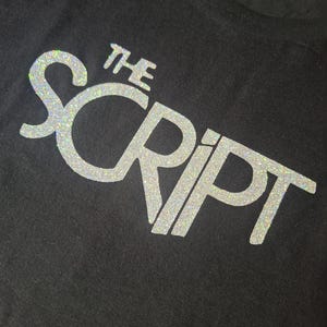 May include: Black t-shirt featuring the band name "The Script" in a shimmering silver font. The text is centrally placed, with "Script" larger than "The". The fabric appears to be a soft cotton material.