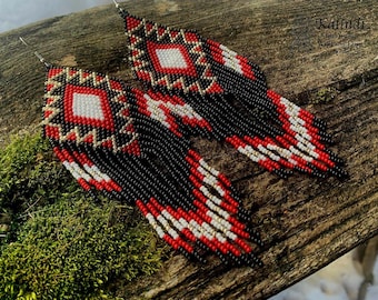May include: Handmade beaded earrings with a geometric design. The earrings feature a central white diamond shape surrounded by red, black, and gold beads. The fringe is black, red, and white. The earrings are displayed on a wooden surface with moss.