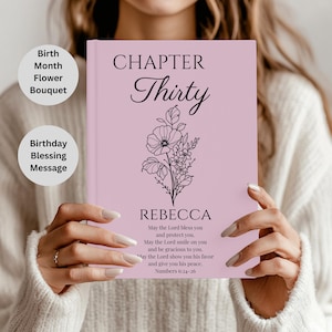 Puede incluir: Un diario rosa claro con las palabras "CHAPTER Thirty" y el nombre "REBECCA" en escritura negra. Una ilustración floral está centrada debajo del texto. El diario también incluye una bendición de Números 6:24-26.