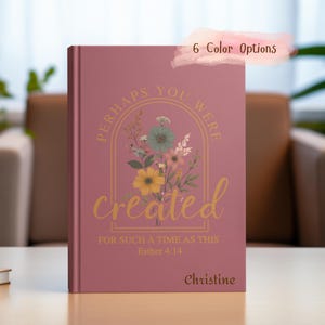Könnte beinhalten: Ein rosa Notizbuch mit einem Blumendesign und dem Text "Perhaps you were created for such a time as this Esther 4:14" in goldenen Buchstaben. Der Name "Christine" ist unten auf dem Notizbuch gedruckt.